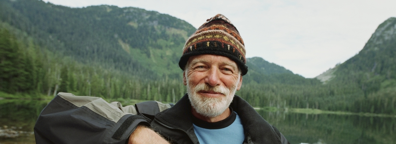 An older gentleman grins in a knitted hat in front of an alpine lake