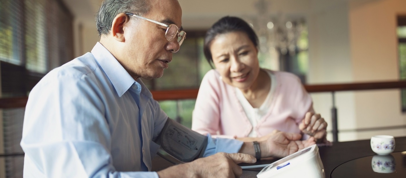 Man checking his blood pressure at home with his wife looking on.