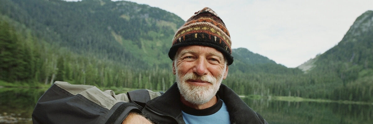 An older gentleman grins in a knitted hat in front of an alpine lake